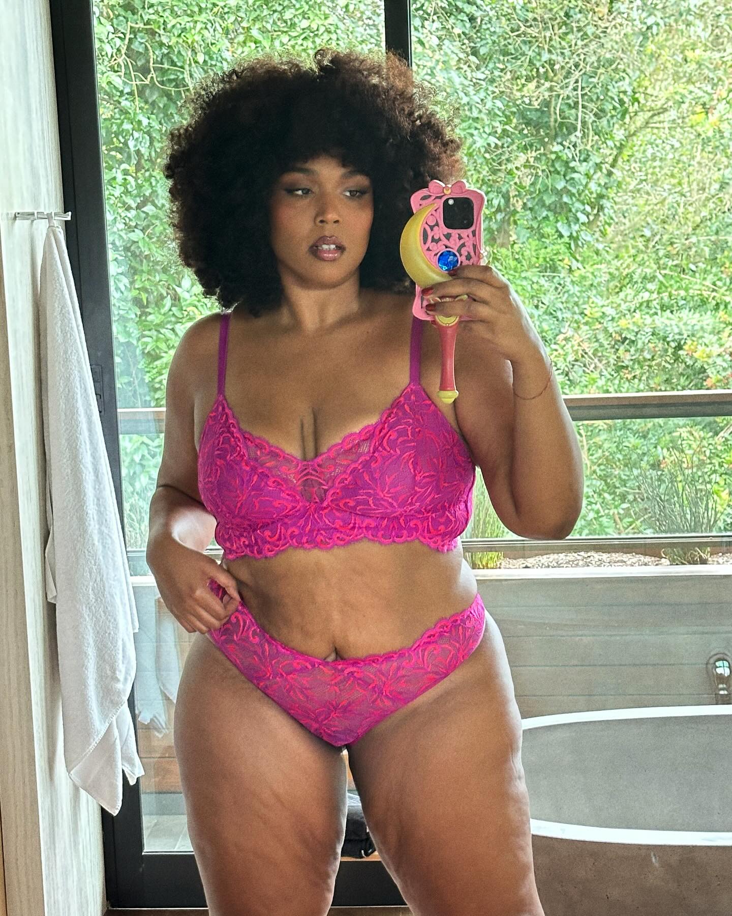 Lizzo in pink lace lingerie, holding a phone with a Sailor Moon case, in a bathroom with a window overlooking greenery.