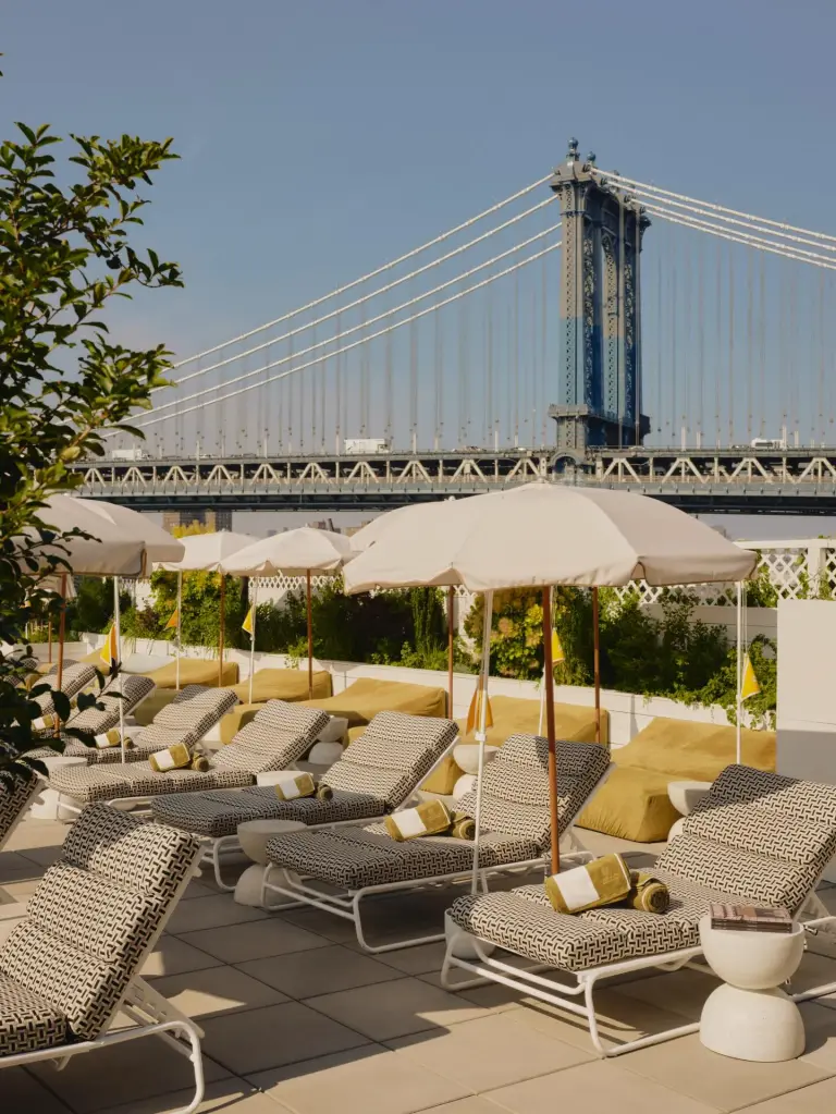 A rooftop lounge area with patterned sun loungers, umbrellas, and the Manhattan Bridge in the background.