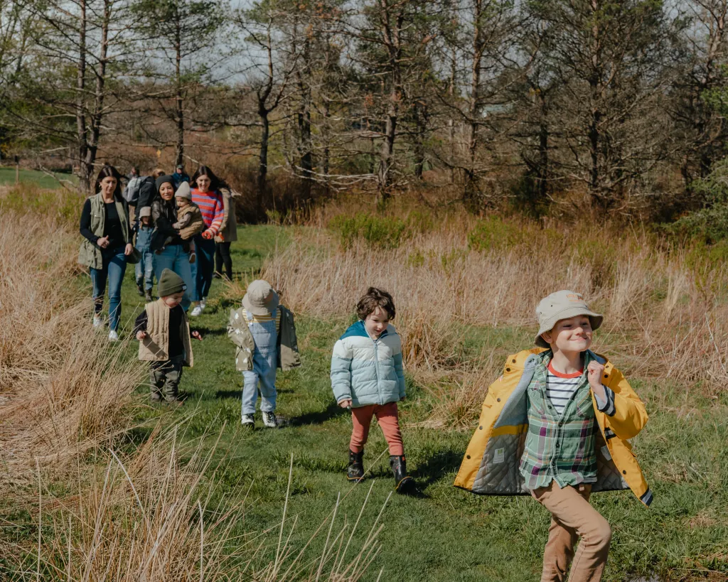 A group of children and adults on a field trip walk along a grassy path surrounded by tall dry grass and trees in the background.
