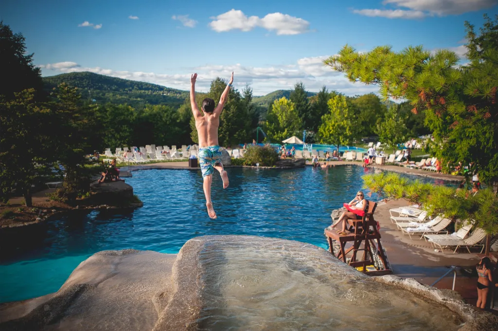 A person in patterned swim trunks jumps into a large outdoor pool at Crystal Springs Resort, with a mountain landscape and blue sky in the background.