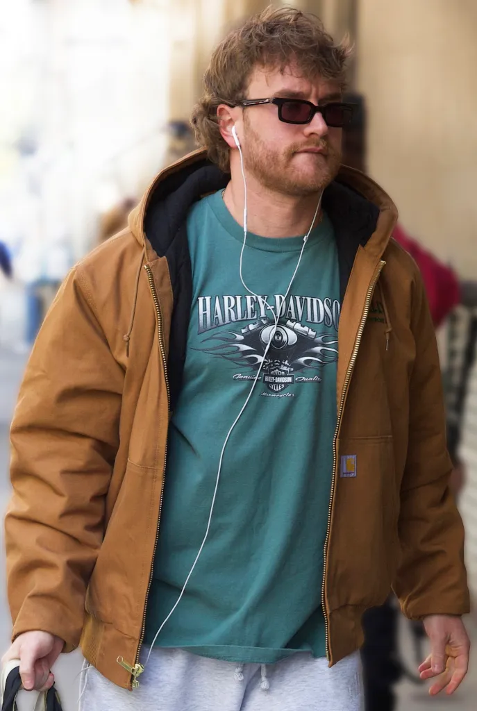 A man in a brown jacket, green Harley-Davidson t-shirt, sunglasses, and earbuds.
