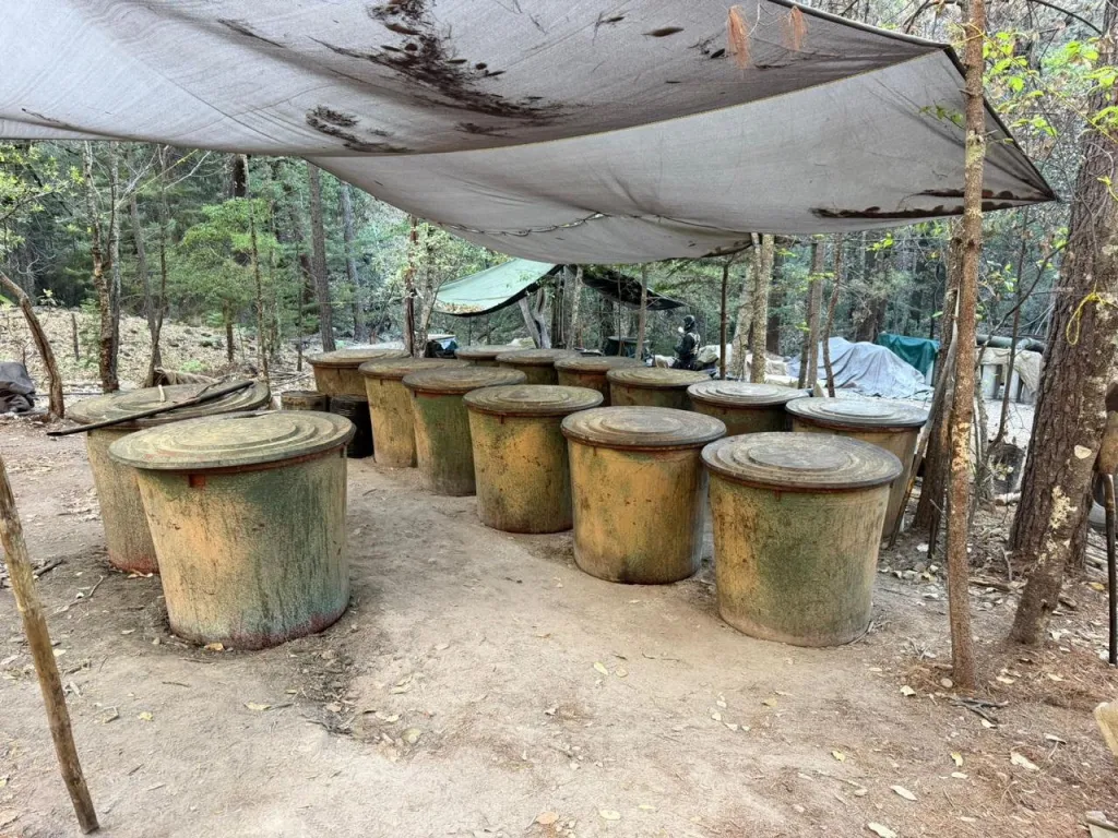 Suspected clandestine methamphetamine lab with large barrels under a tarp in a wooded area.