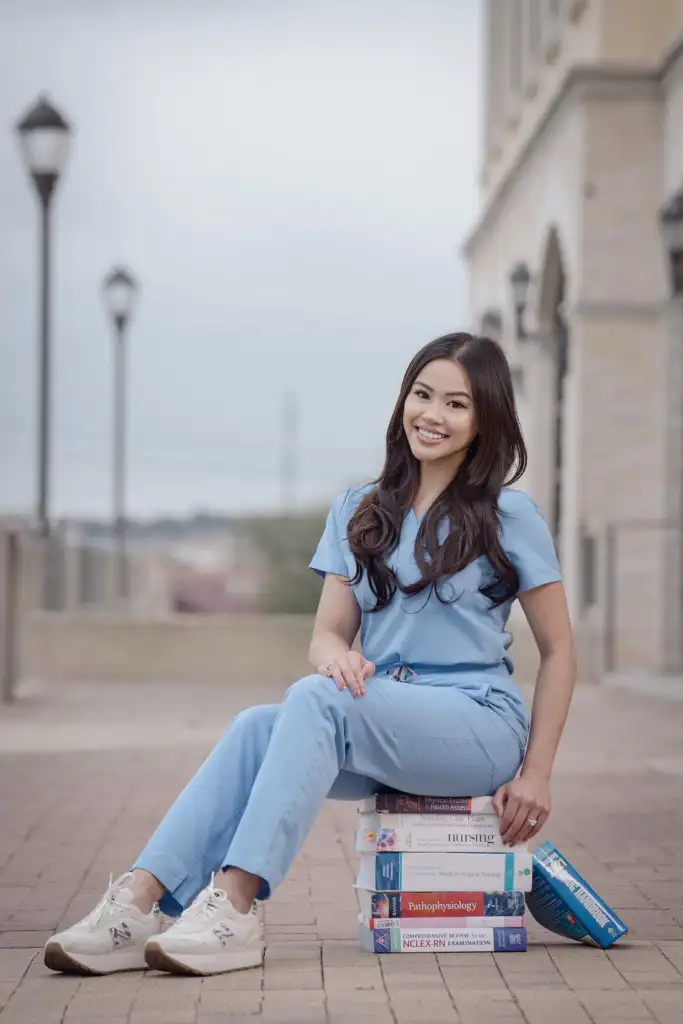 A smiling nurse in light blue scrubs sits on a stack of nursing textbooks.