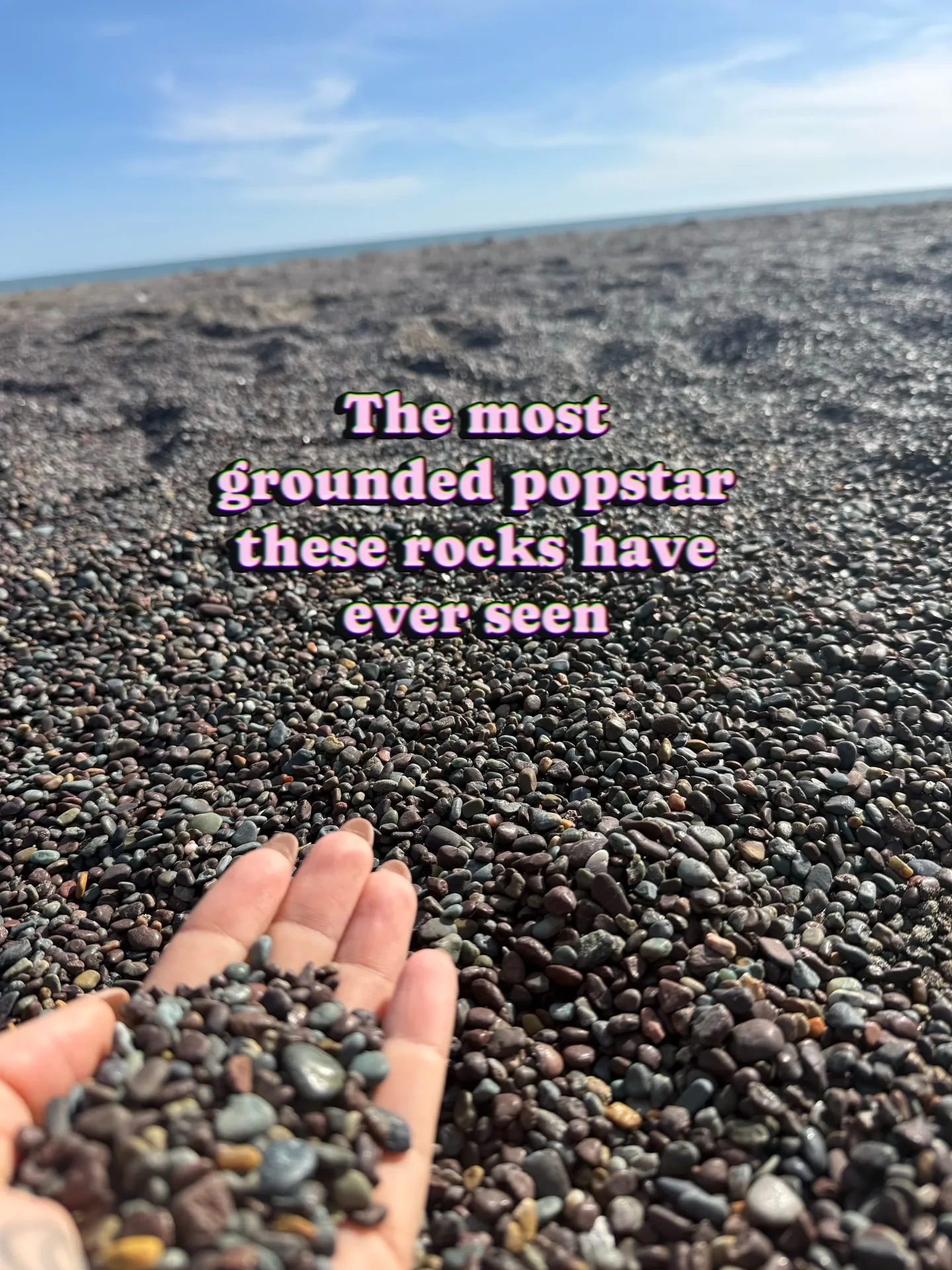 A hand holding small, dark pebbles on a rocky beach with the ocean in the background.