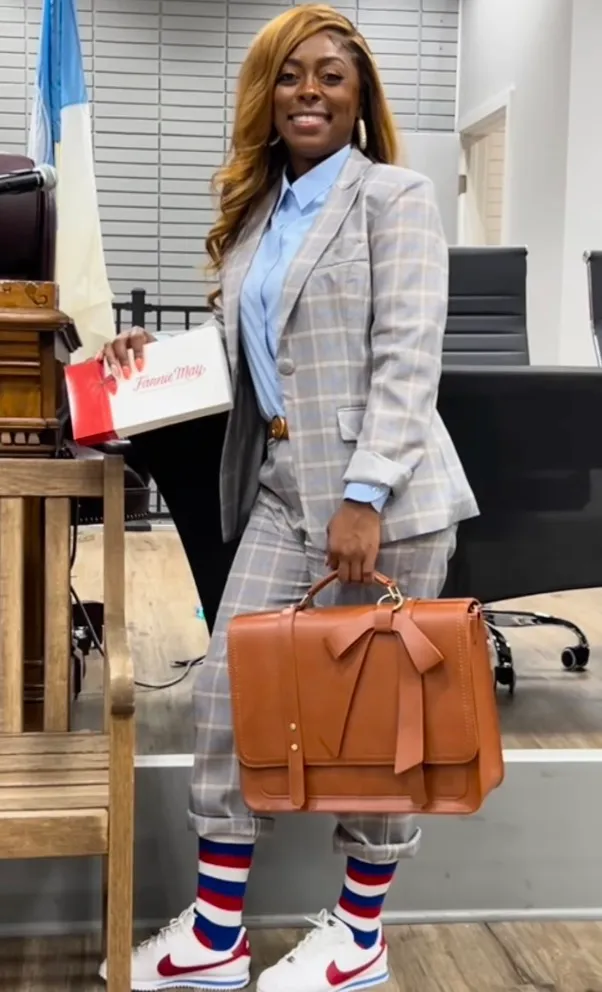 Tiffany Henyard, Mayor of Dolton, Illinois, in a gray plaid pantsuit and sneakers, holding a Fannie May box and a brown briefcase.