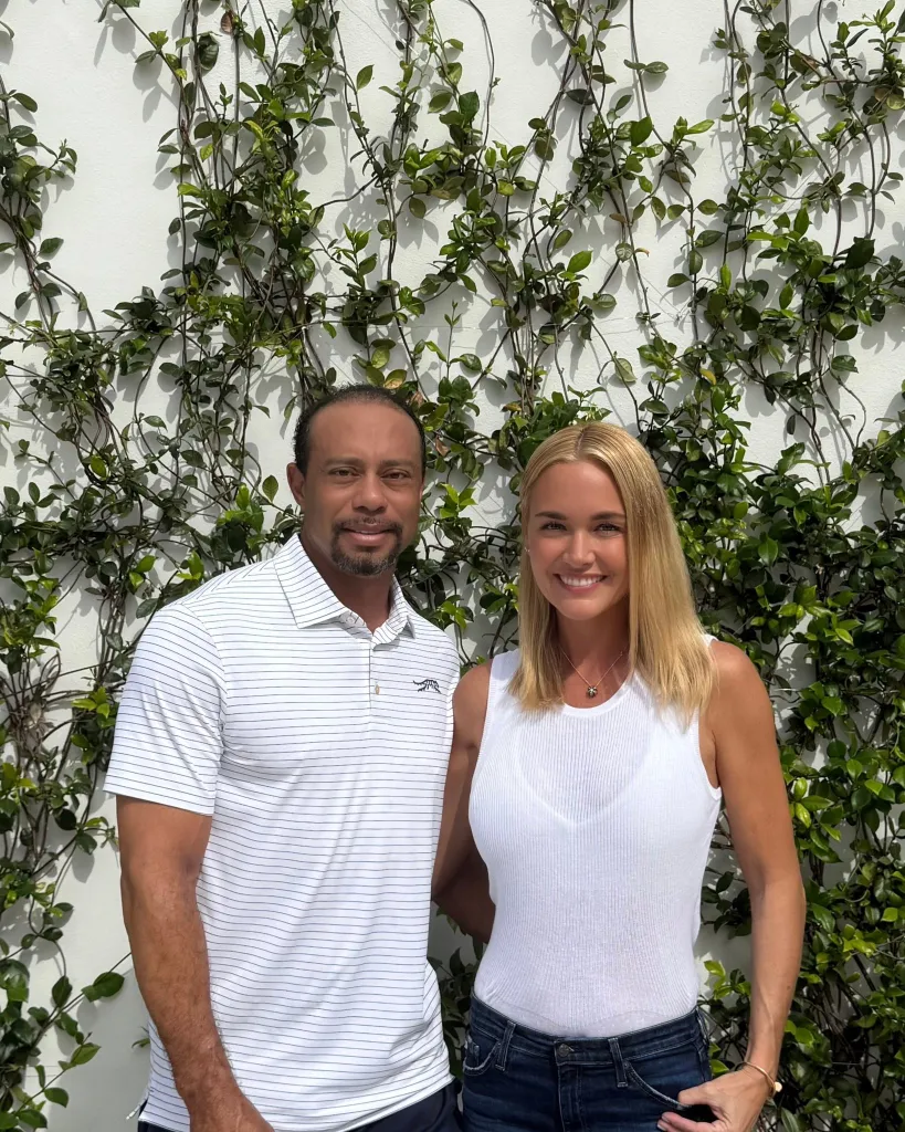 Tiger Woods and a blonde woman smiling in front of an ivy-covered wall.