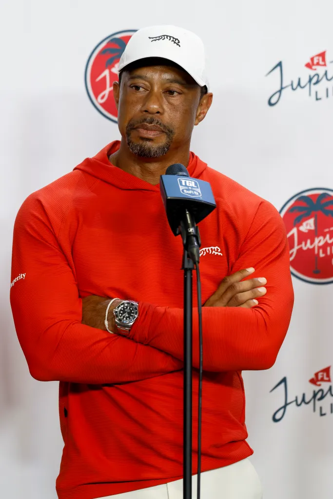 Tiger Woods speaks at a press conference, wearing a white cap, red hooded shirt, and a wristwatch, with a microphone in front of him.