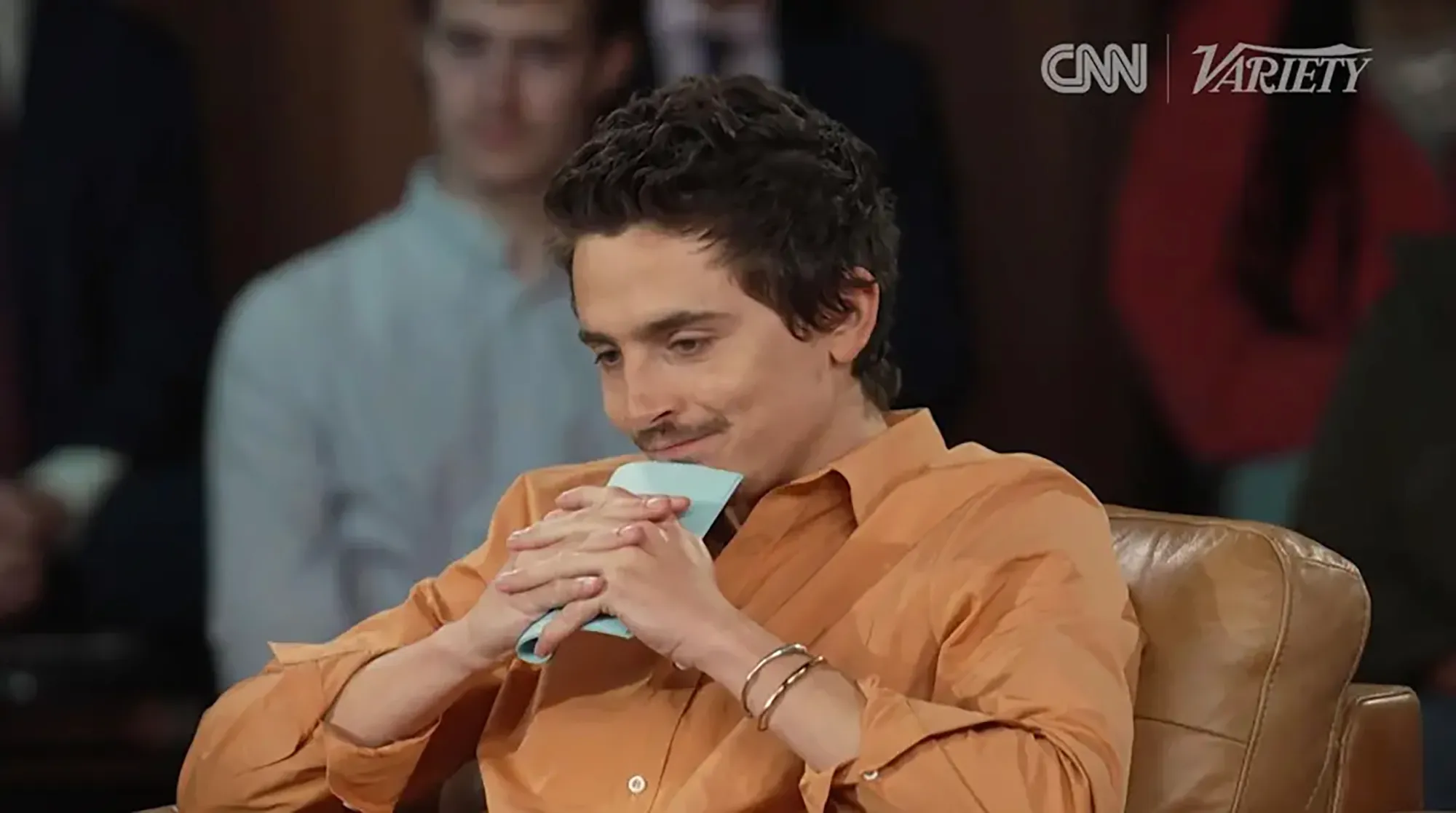 Timothée Chalamet, in an orange shirt, seated with clasped hands and a blue napkin, looking down and to the left.