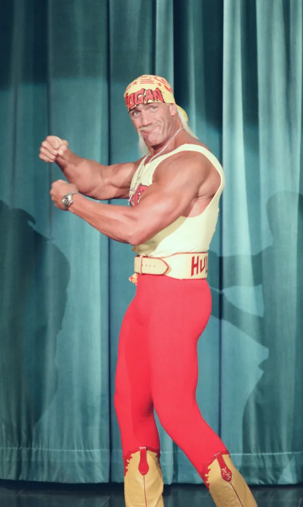 Hulk Hogan flexing his biceps while wearing a yellow tank top, red pants, and yellow bandana.