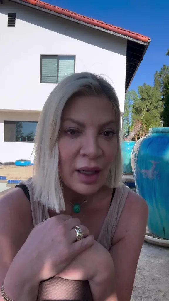 Tori Spelling, with shoulder-length blonde hair and a green necklace, speaking outdoors.