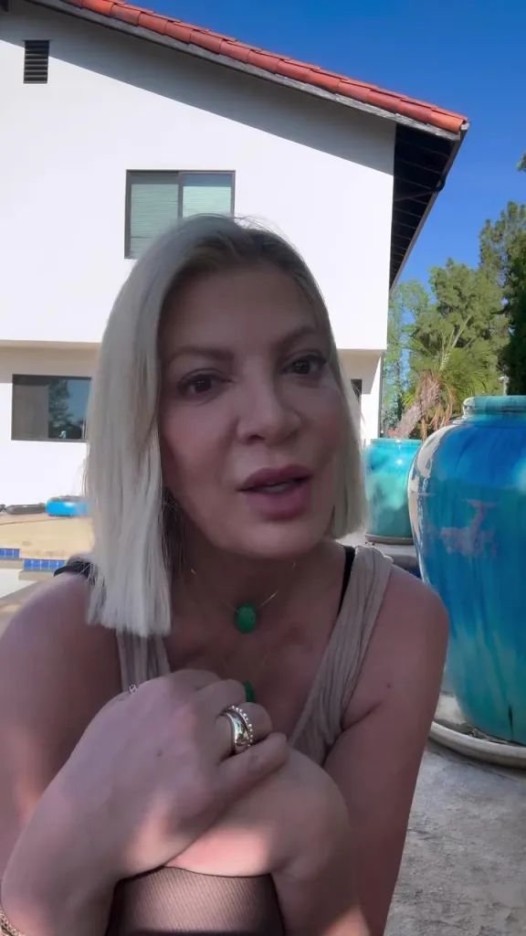 Tori Spelling speaking to the camera in front of a white house with a blue pot next to a pool.