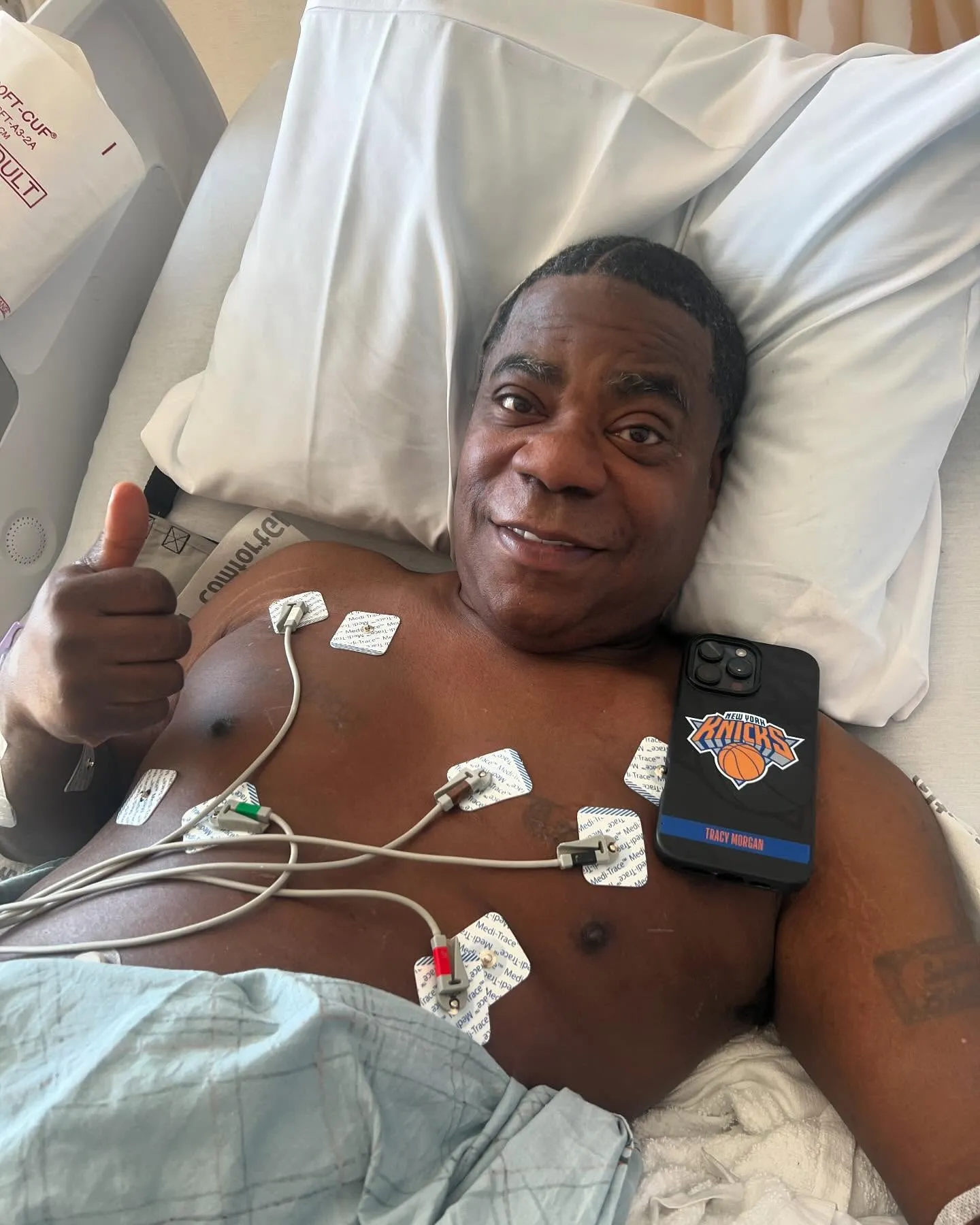 Tracy Morgan giving a thumbs up in a hospital bed with medical sensors on his chest.