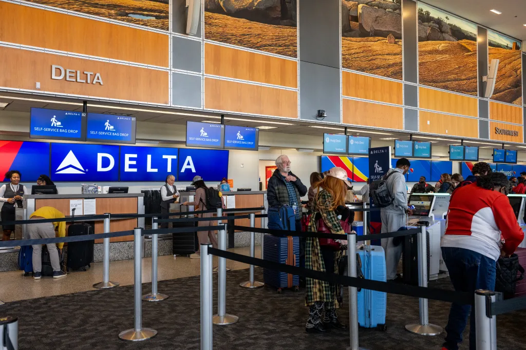 Travelers wait in line at Delta Airlines check-in and self-service bag drop counters at an airport.