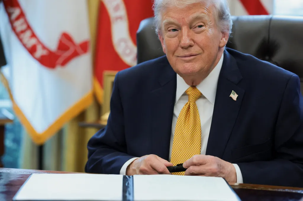 U.S. President Donald Trump seated at a desk with a pen in his hands, smiling, during an event to sign an executive order in the Oval Office.