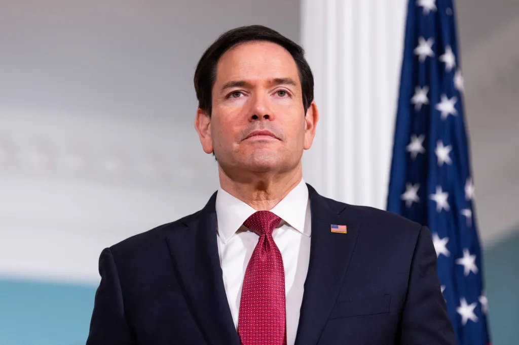 U.S. Secretary of State Marco Rubio wearing a suit, red tie, and American flag pin.