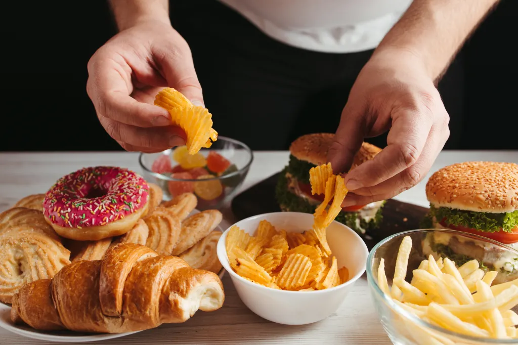 A person's hands reaching for potato chips from a table laden with various junk foods like donuts, croissants, burgers, and french fries.