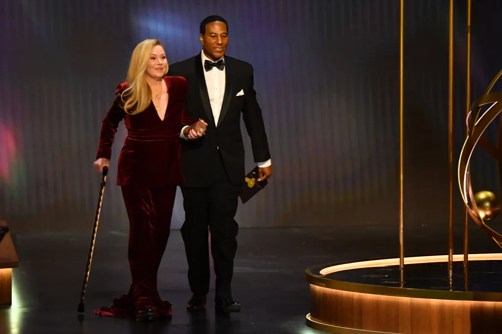 Christina Applegate, in a red velvet suit and holding a cane, is escorted by a man in a tuxedo onto a stage.