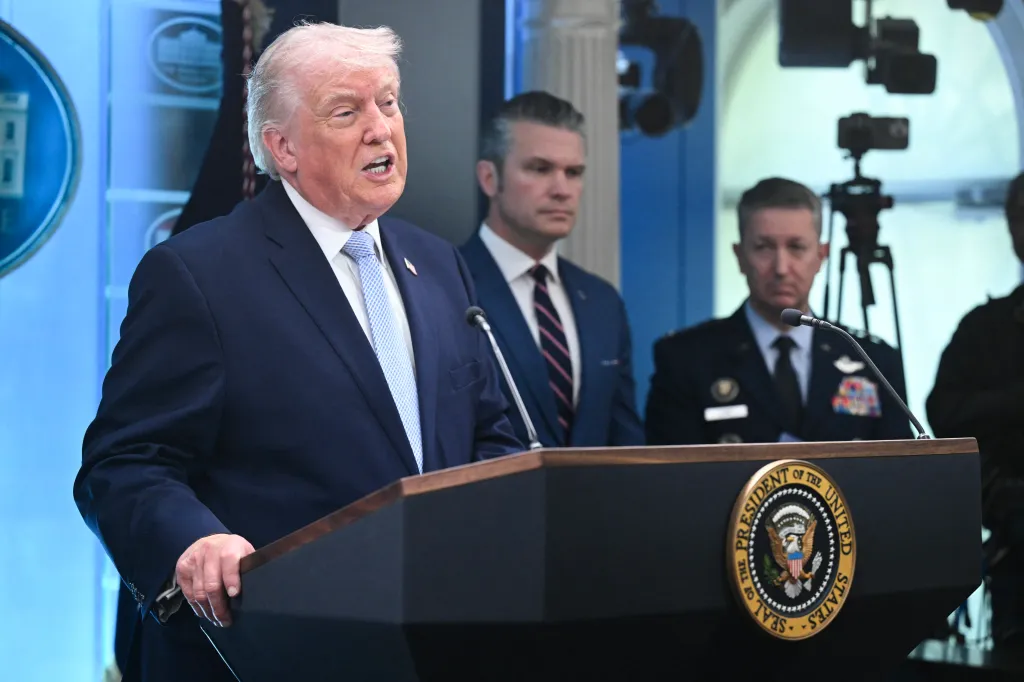 Donald Trump speaking at a podium with two men in suits standing behind him.