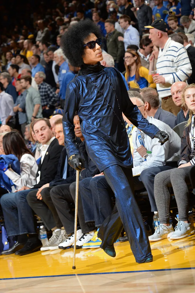 Prince walking with a cane in a metallic blue outfit and sunglasses.