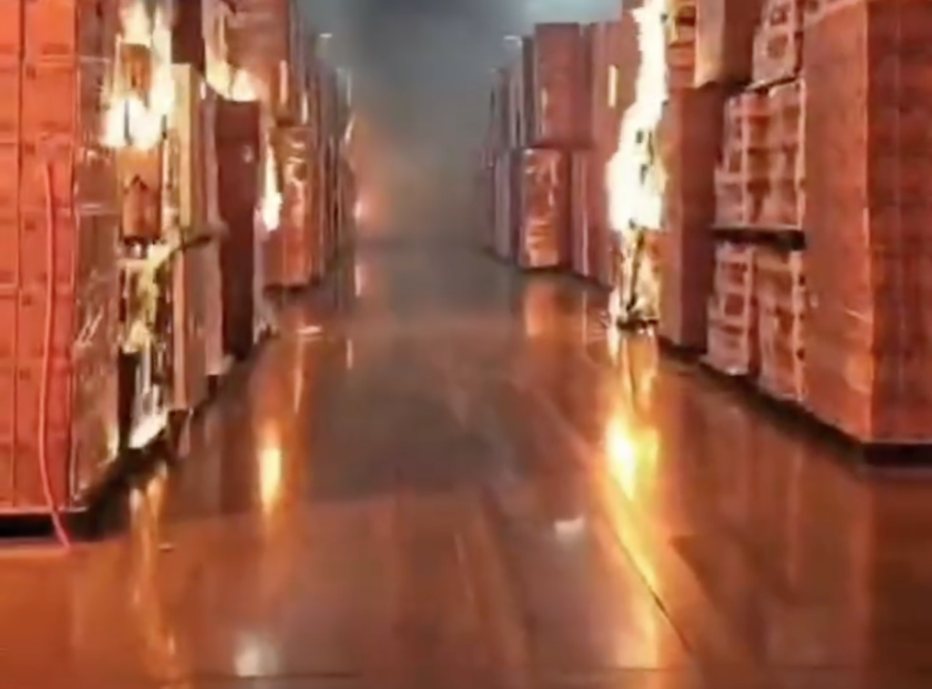 Video still of a warehouse aisle where boxes are on fire and flames are reflecting on the floor.