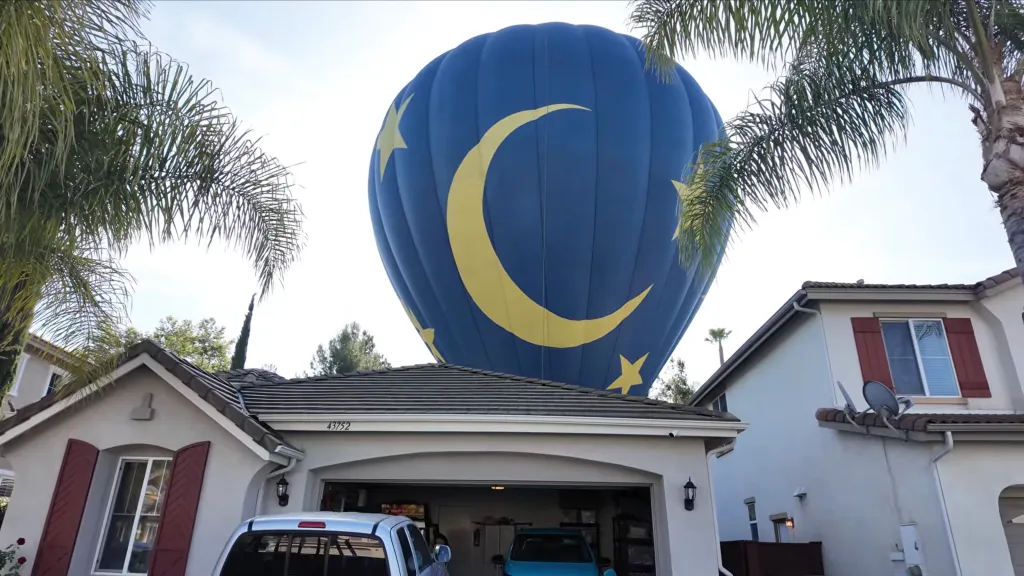 A blue hot air balloon with yellow stars and crescent moon deflates above two houses.