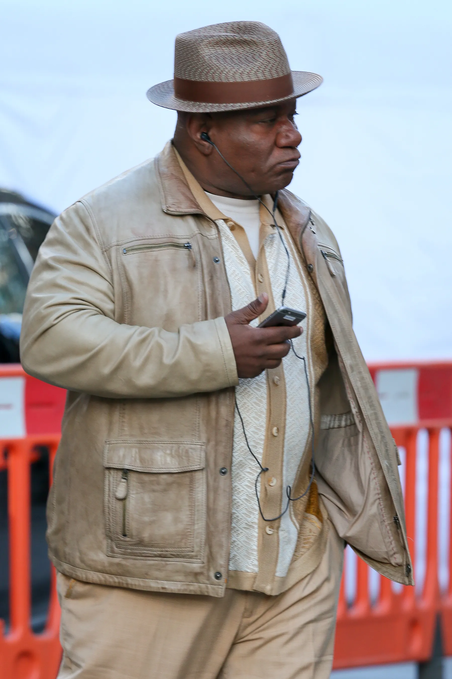 Ving Rhames in a light brown fedora, matching leather jacket, and patterned shirt, holding a phone with wired earbuds in.