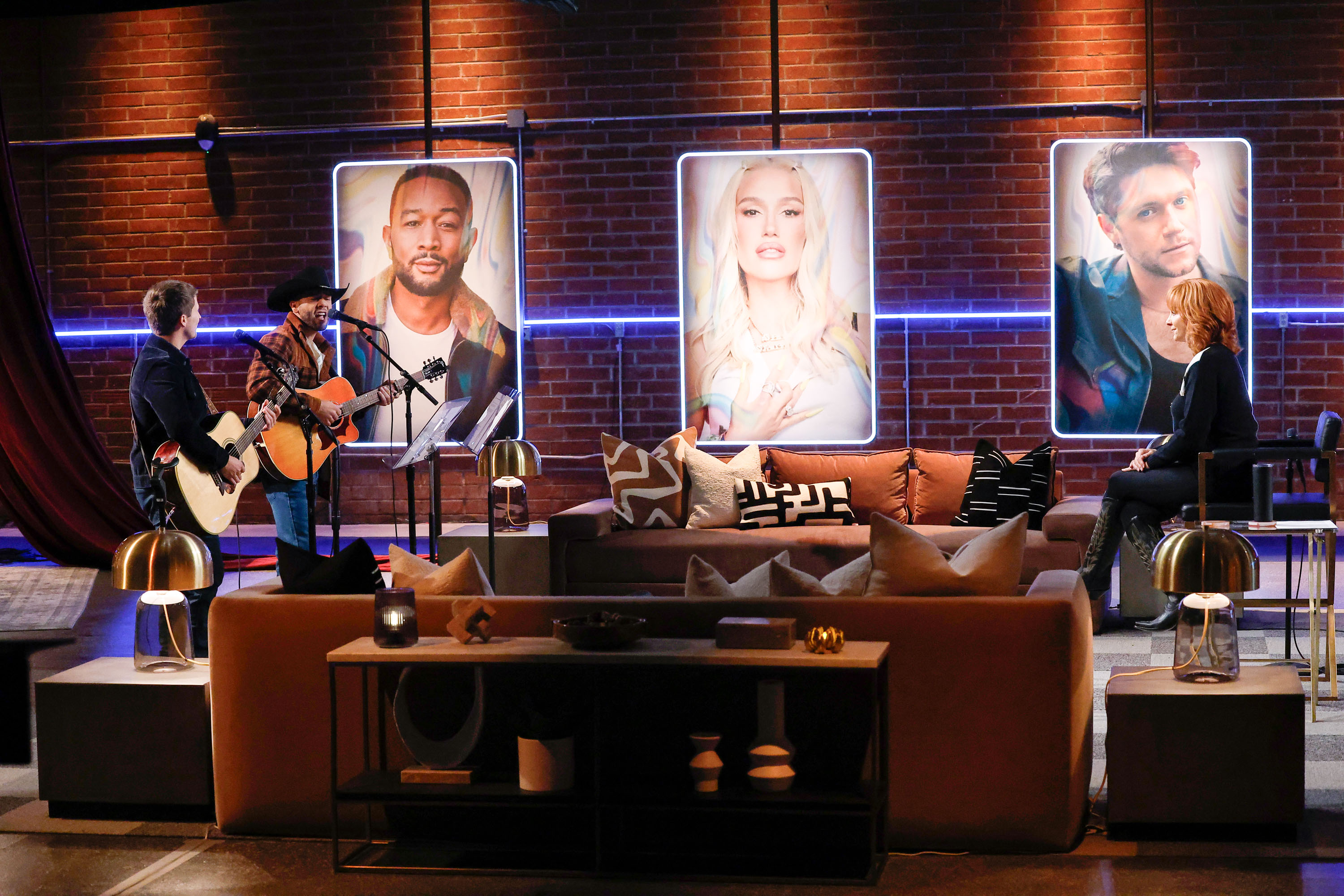 Two men sing and play guitars on stage in front of portraits of John Legend, Gwen Stefani, and Niall Horan, while Reba McEntire watches from a chair.