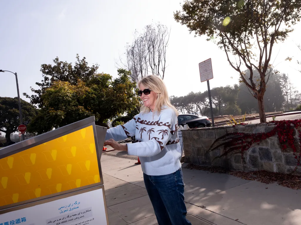 A blonde woman in sunglasses and a light blue sweater casts her ballot into an official drop box in Pacific Palisades.