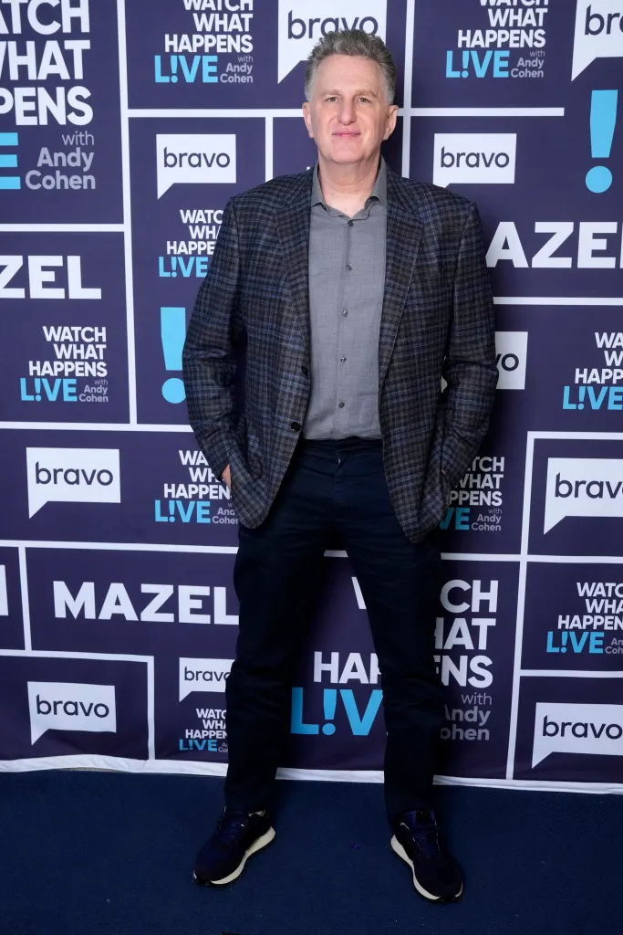 Michael Rapaport, in a grey checked blazer, grey shirt and black pants, standing in front of a