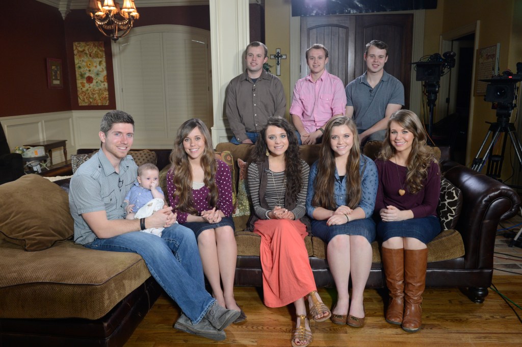 The Duggar children and spouses, with one holding a baby, seated on a sofa.