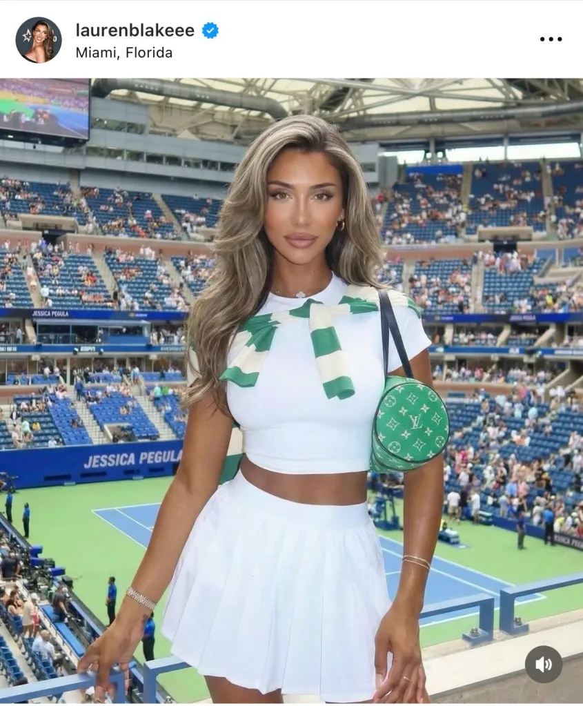 Lauren Blake Boultier at a tennis stadium, wearing a white crop top and skirt, with a green and white striped sweater over her shoulders and a green Louis Vuitton bag.