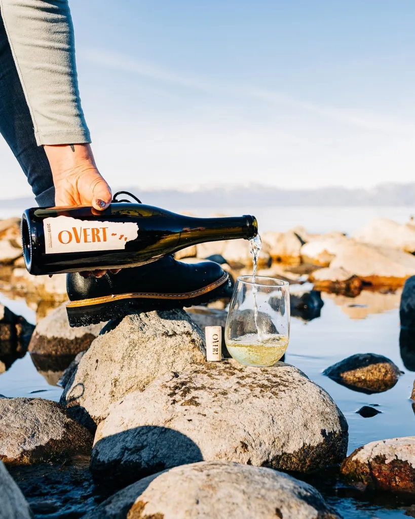 A hand pouring Overt wine from a bottle into a stemless glass, set on rocks next to water.