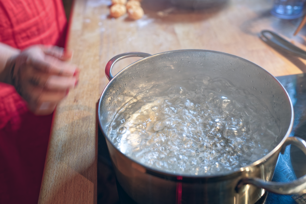 A pot of boiling water on a stove with a person's hands in a red apron preparing gnocchi in the background.