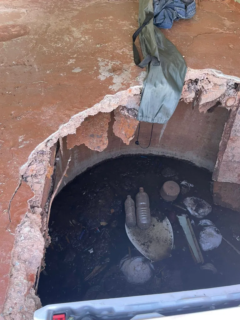 Broken concrete floor revealing a deep, dark pit filled with dirty water, bottles, and other debris.