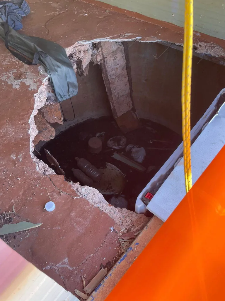 A large hole in a red concrete floor reveals a pit filled with debris and dark liquid.