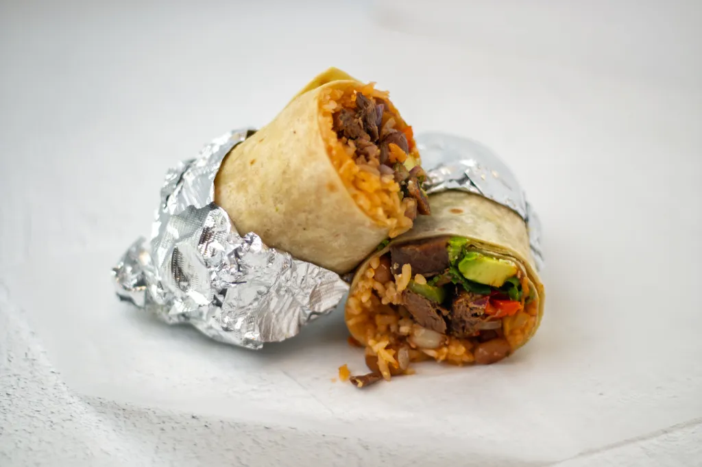 Two burritos, wrapped in foil and cut in half, filled with rice, meat, avocado, and vegetables.