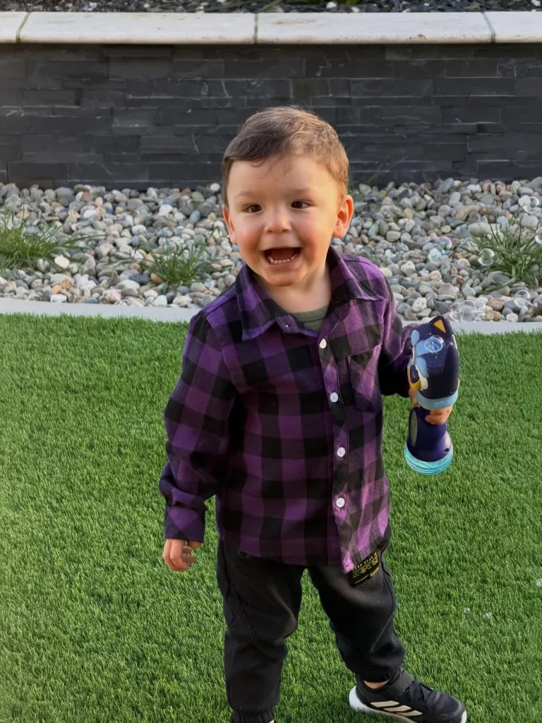 Baby Jaxon in a purple and black plaid shirt, holding a bubble blower, standing on grass.