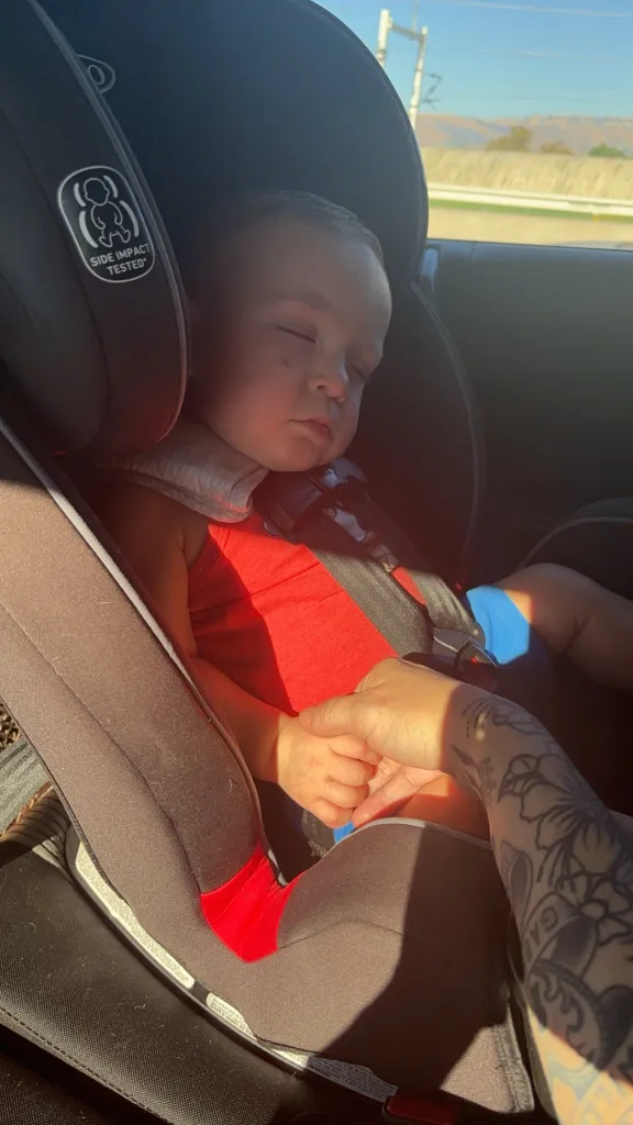 Baby Jaxon asleep in a car seat, holding an adult's hand.