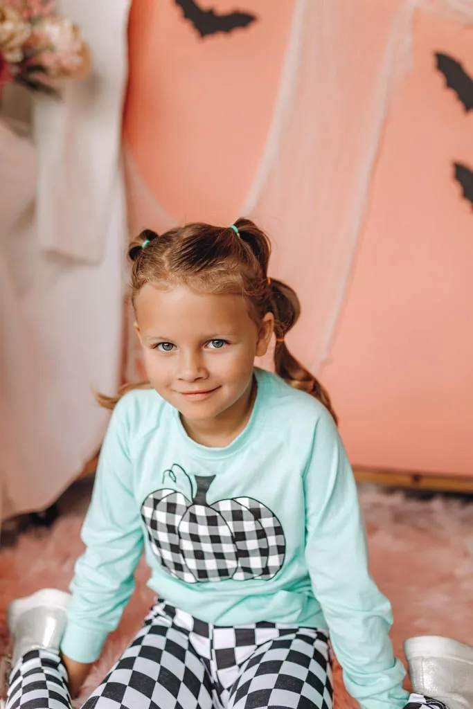 A young girl with blonde hair in pigtails smiles, wearing a light blue shirt with a plaid pumpkin design and black and white checkered pants.