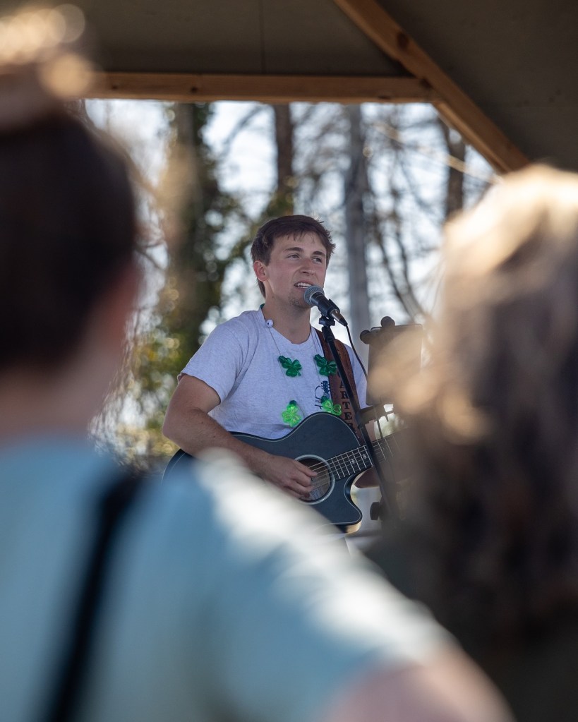 Dylan Carter performing with a microphone and guitar, wearing a white shirt and a shamrock necklace.