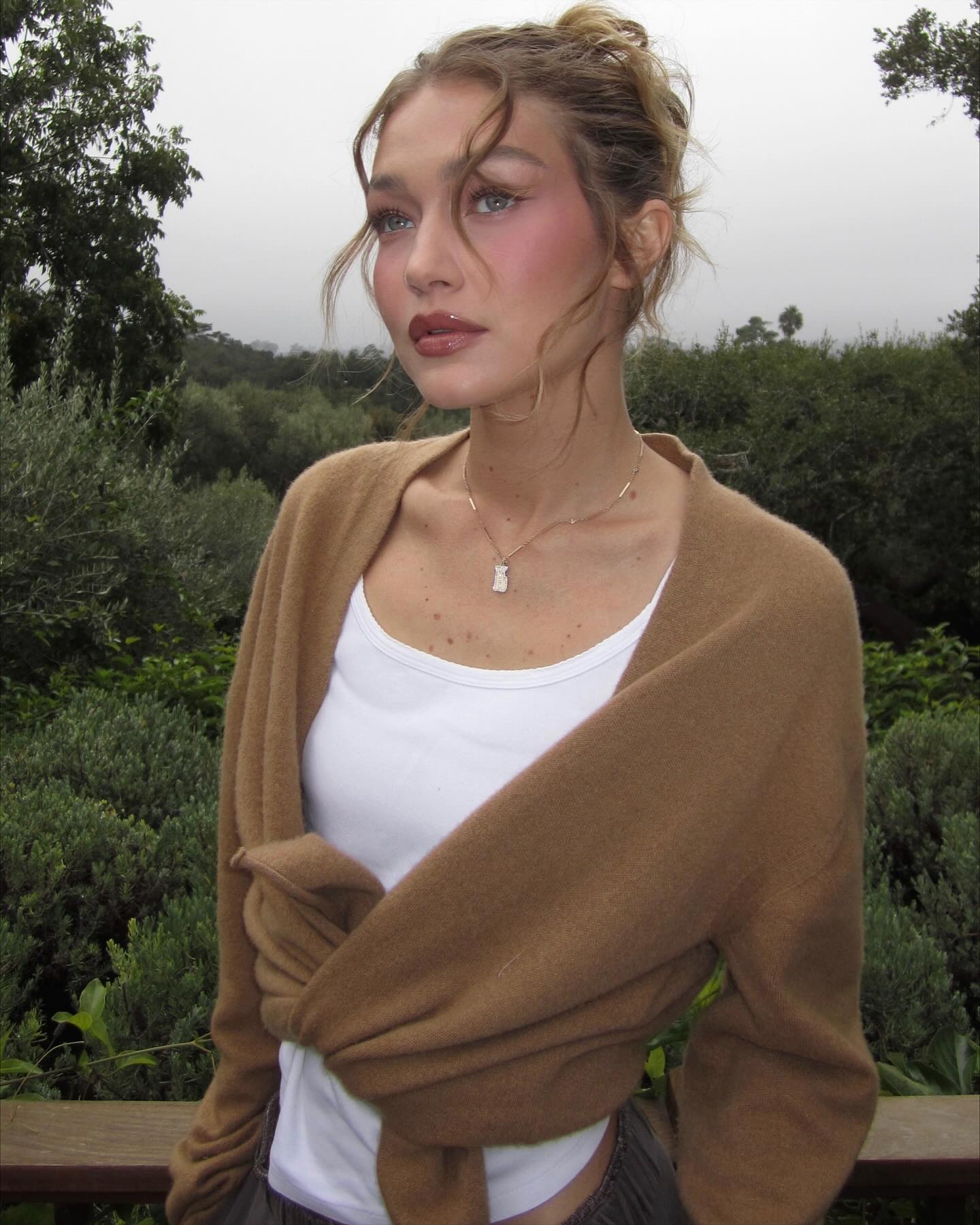 Gigi Hadid wearing a white top and a brown sweater, looking to the left.