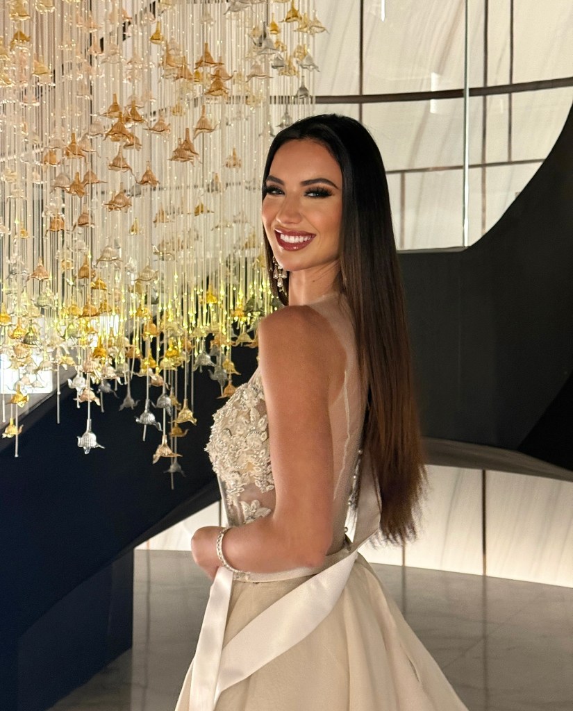 Melanie Shiraz Asor in a light dress with intricate beading, smiling towards the camera, with a modern chandelier behind her.