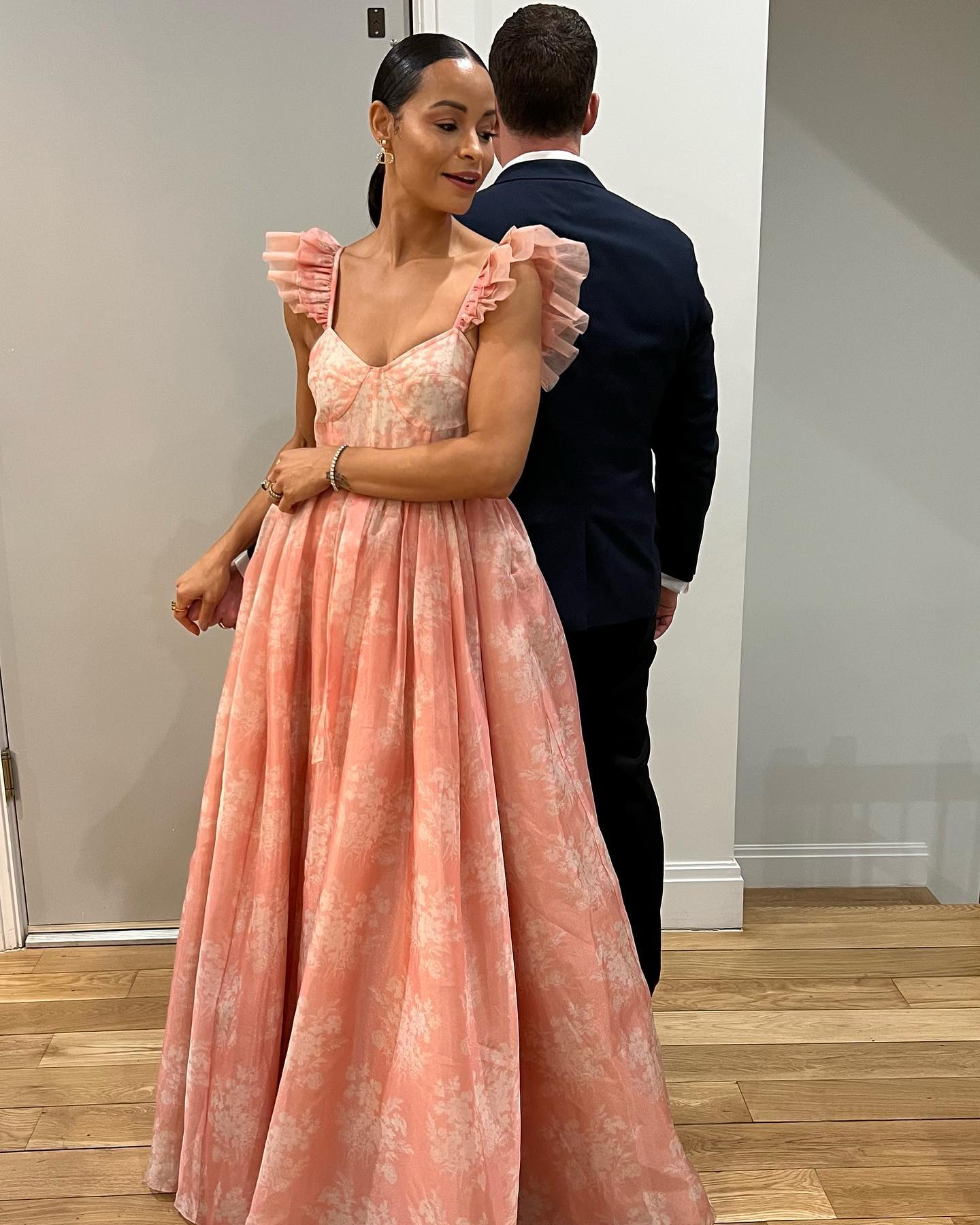 A woman in a floral peach dress with ruffled sleeves poses next to a man in a suit, seen from behind.