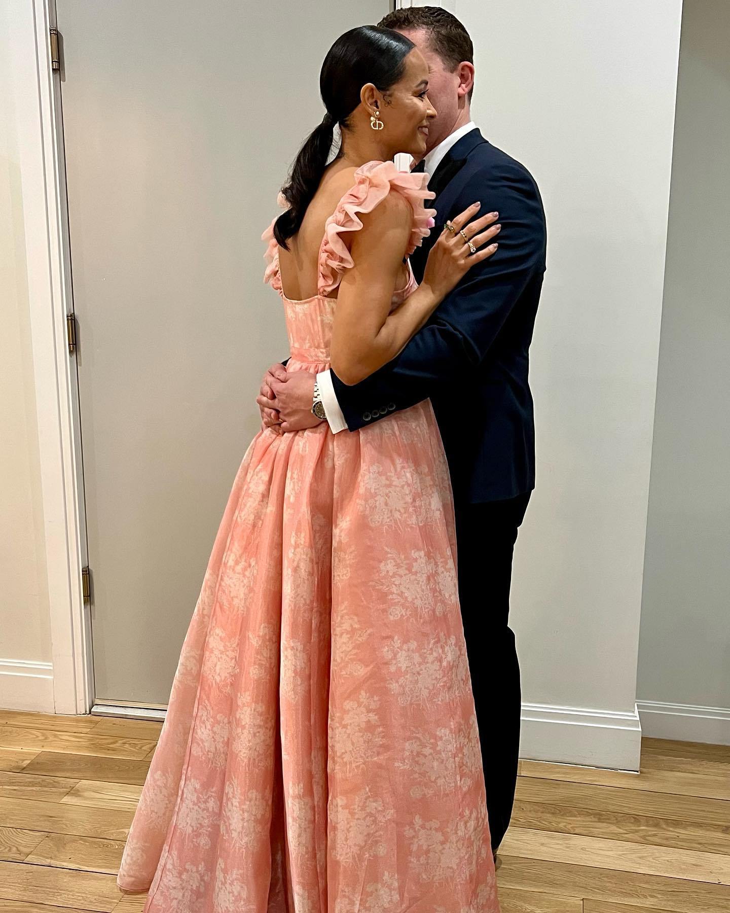 Sai De Silva in a pink floral gown with ruffled shoulders embracing a man in a dark suit.