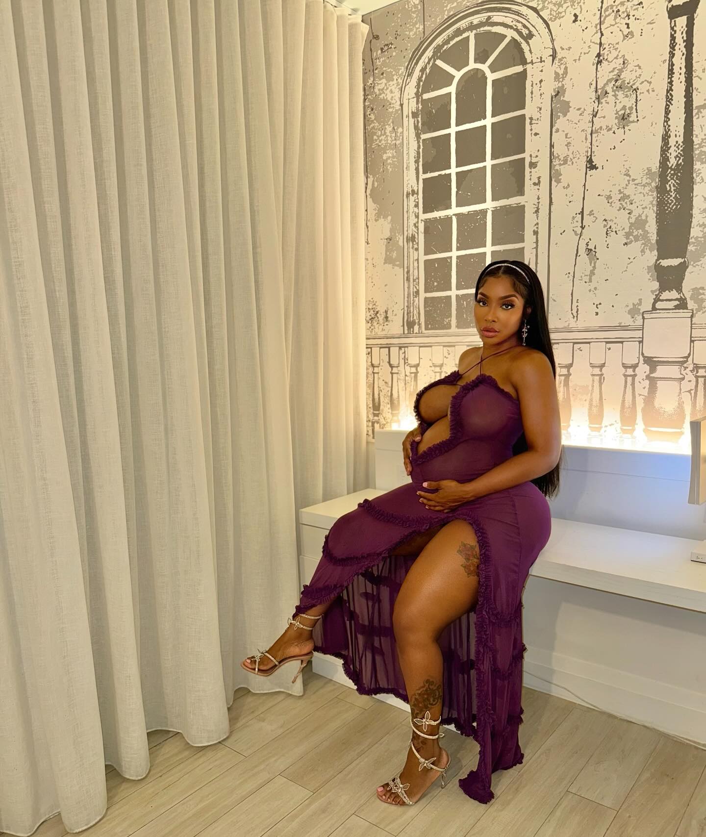 Ayesha Howard in a long, sheer purple dress while cradling her baby bump.
