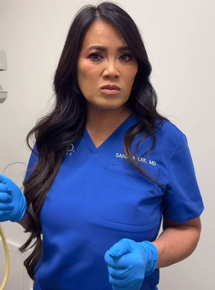 Dr. Sandra Lee, also known as Dr. Pimple Popper, wearing blue scrubs and blue gloves, looking serious.