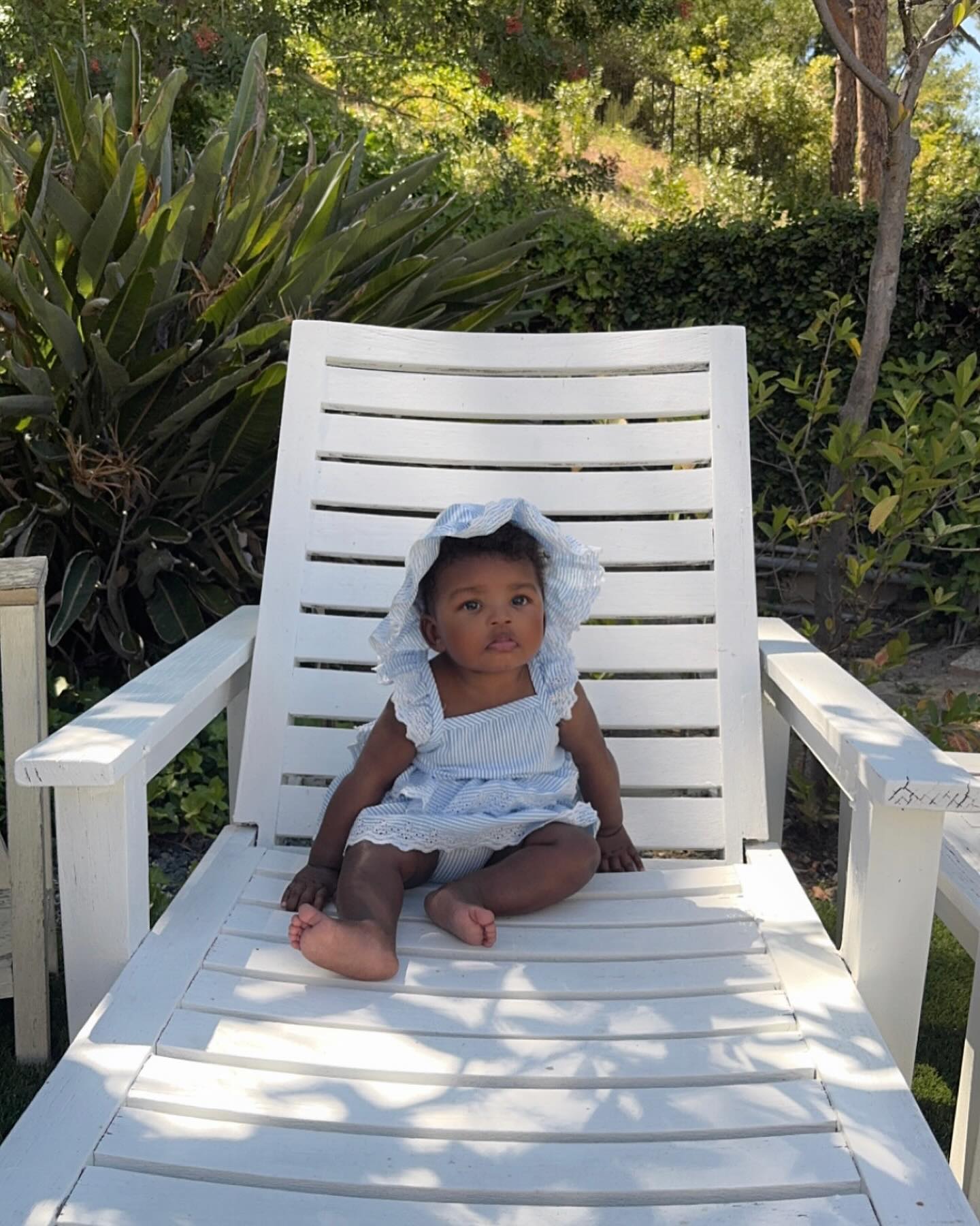 A baby, Aubri, sitting in a white wooden chair outdoors.