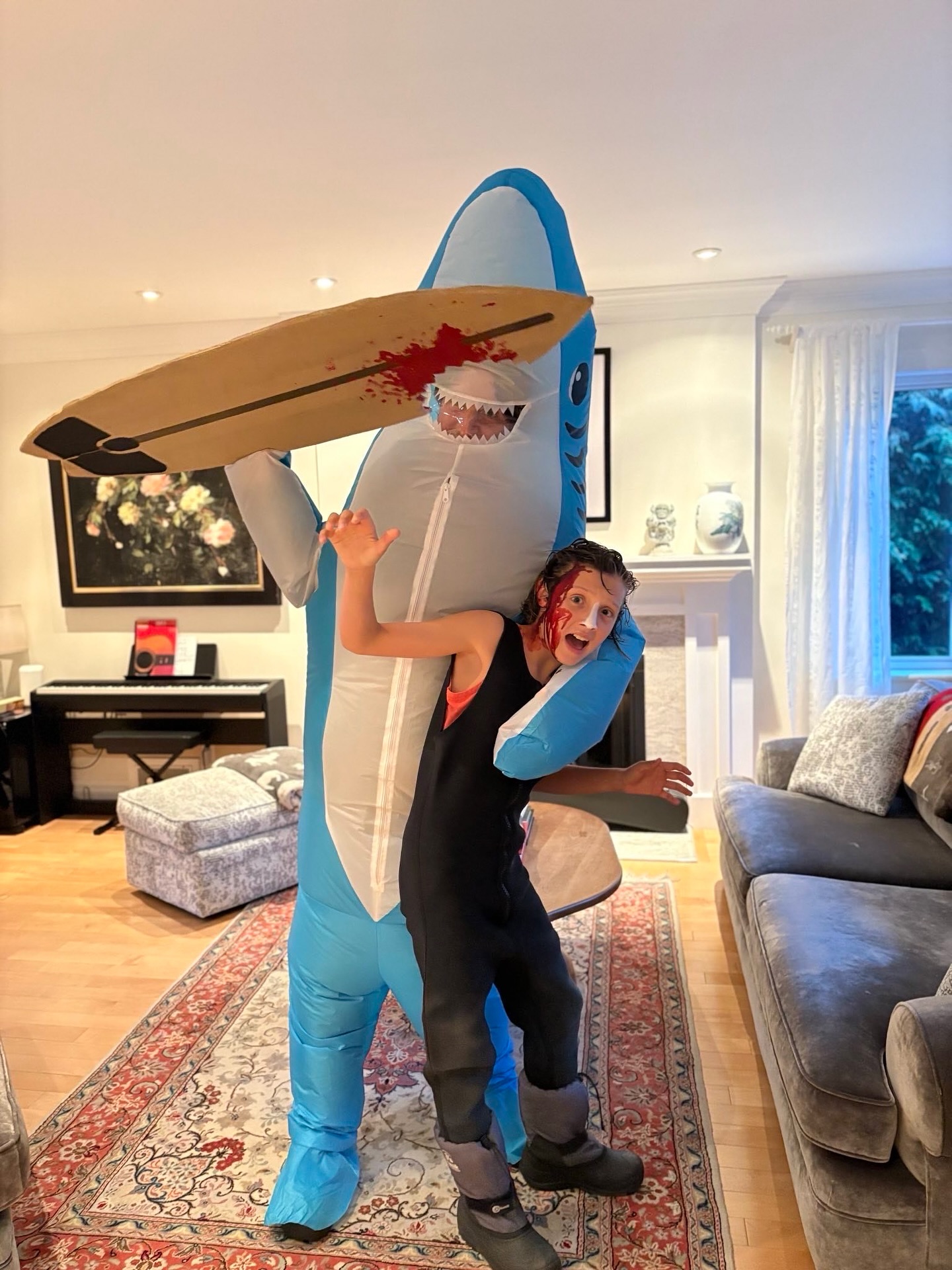 Person in shark costume carrying a surfboard with fake blood, holding another person dressed as a surfer with fake blood on their face.