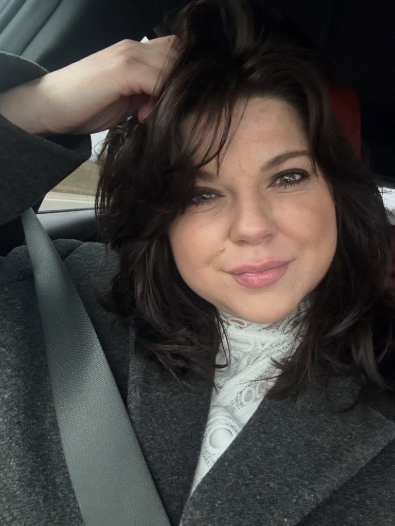 Amy Duggar in a car wearing a white lace top and gray coat.