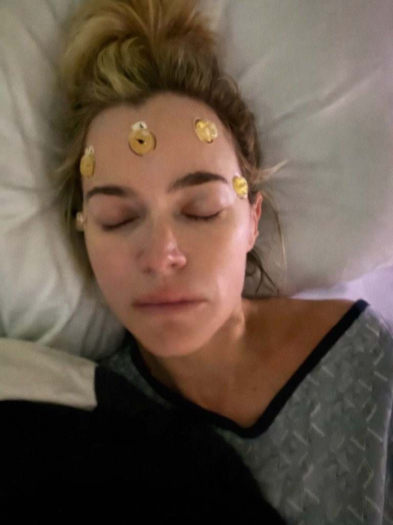Teddi Mellencamp in a hospital gown, with medical electrodes on her forehead, lying in bed with her eyes closed.